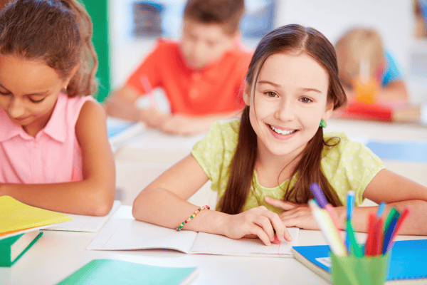Happy schoolgirl in classroom with classmates and colourful stationery.