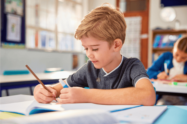 Young boy focused on writing in classroom during school readiness assessment.